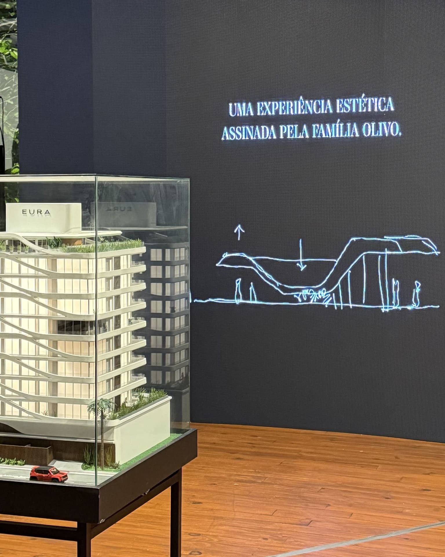 Telão do lançamento do Eura One com a frase "Uma experiência estética assinada pela Família Olivo" e um croqui, ao lado da maquete física do prédio