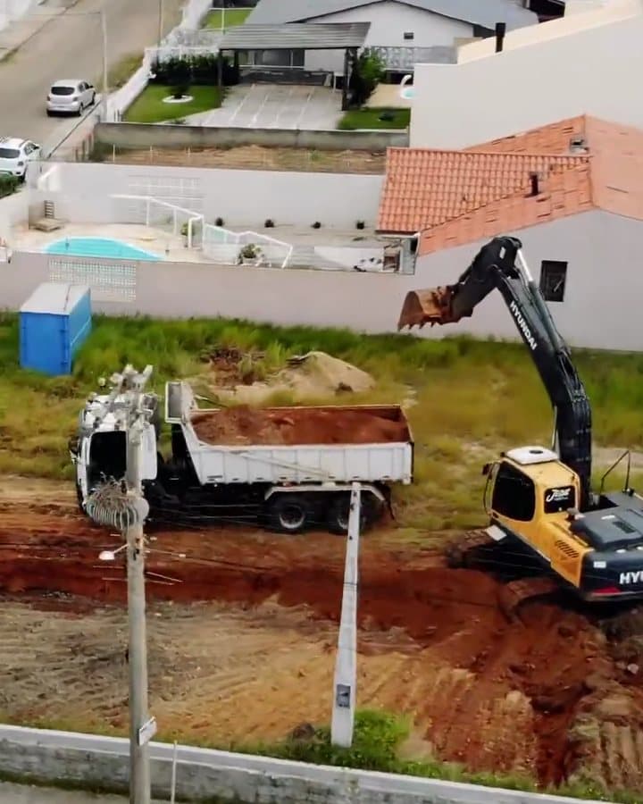 Vista aérea do terreno no início da obra, com uma escavadeira e um caminhão removendo terra